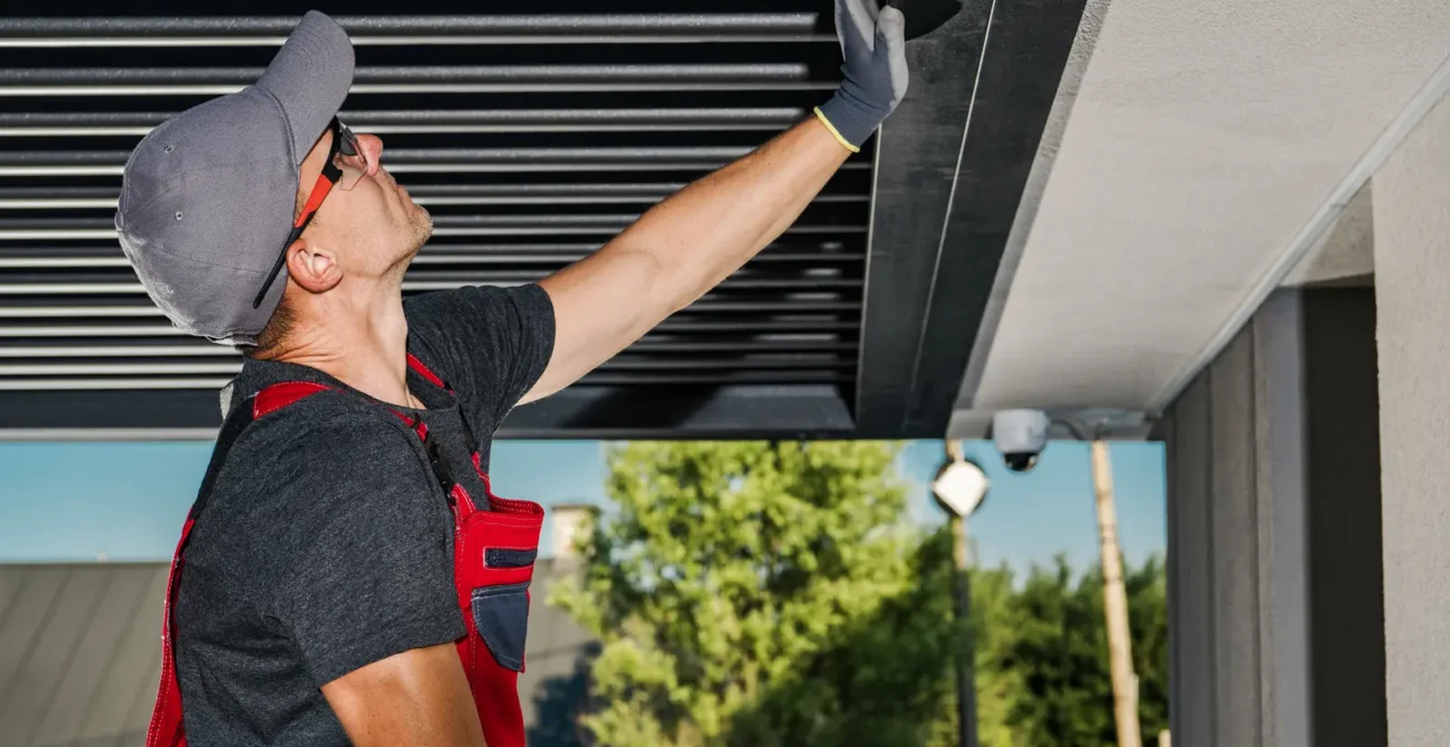 Un hombre limpia e inspecciona las lamas y canalones de una pérgola bioclimática de aluminio desde una escalera.